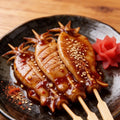 Glazed squid skewers fanned on a dark plate with a shiso leaf and pickled garnish
